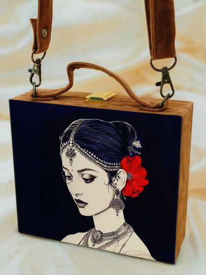 This suitcase-style bag offers a sophisticated and artistic aesthetic, featuring a portrait of a traditional Indian woman. The contrast of the dark background makes the details of her jewelry and the red flower pop.