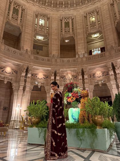 A side profile shot at Umaid Bhawan Palace, showcasing a luxurious maroon velvet saree against the grand, historic architecture.