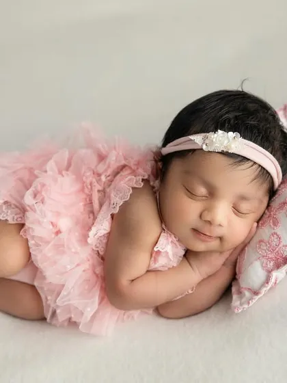 A sweet baby girl in a pink tutu romper, resting her head on a small floral pillow. The white background keeps the image bright and airy.
