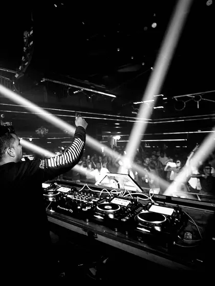 A black and white photo capturing the scale of the venue and the crowd. The spotlights create a dramatic effect, highlighting the connection between the DJ and the audience.