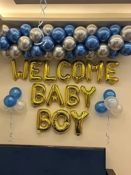 A classic and cheerful 'Welcome Baby Boy' setup. This features bold gold foil letter balloons against a wall, topped with a garland of metallic blue and silver balloons to celebrate his arrival.