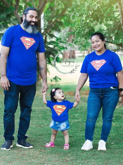 A super family. Mom, dad, and their little girl are all wearing matching Superman t-shirts for a fun and playful family portrait in the park.