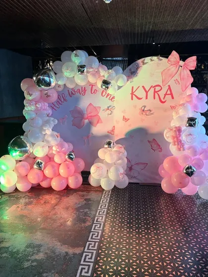 A "Half way to one" celebration for baby Kyra. The backdrop is decorated with a pink and white balloon heart, silver disco balls, and a butterfly and bow theme.