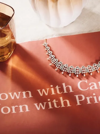 A sophisticated flat lay featuring a diamond necklace on a branded card with the tagline "Grown with Care, Worn with Pride." The warm lighting and the glass of whiskey nearby create a mood of quiet luxury and confidence.