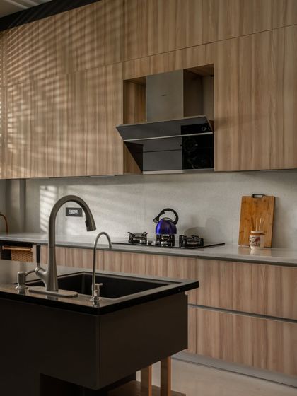 The kitchen in Twin Villa features clean lines and a warm wood finish. The play of light and shadow from the blinds adds a dynamic quality to the minimalist design, highlighting the texture of the materials.