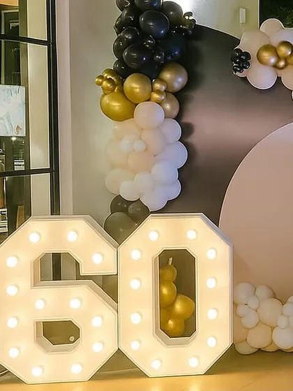 A close-up of large, illuminated '60' marquee numbers, a key element for milestone birthday parties. The numbers are set against a stylish black, gold, and white balloon backdrop.