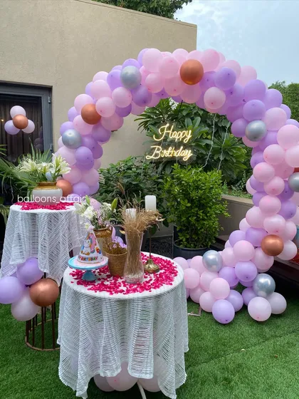 A lovely outdoor birthday party with a pastel theme. The decor features a balloon arch in shades of pink, purple, and rose gold, with two cake tables.