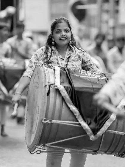 A powerful black and white shot of a female dhol player, her strength and determination evident as she carries the large drum.