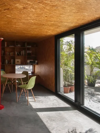 The interior of a compact workspace designed for a journalist. Large glass panels flood the space with natural light, while the use of oriented strand board (OSB) on the walls and ceiling creates a warm, textured, and cost-effective environment.