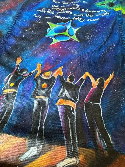A detail shot from the back of the black Coldplay jacket, focusing on the painted figures and the galaxy art. This design includes lyrics from "Adventure of a Lifetime" and "Diamonds."