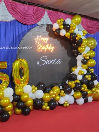We designed this complete stage setup for a 50th birthday party in a hall. It features a custom circular backdrop with the name 'Sweta', a neon sign, large foil '50' balloons, and an extensive balloon garland in black, gold, and white.