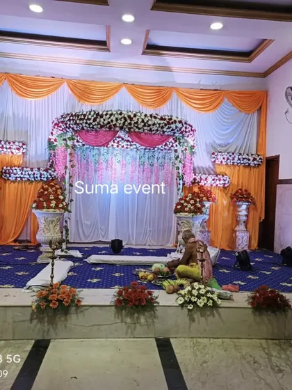 This is a classic flower decoration for a wedding or engagement ceremony in a smaller hall. The setup includes a floral arch over the main seating area, matching flower arrangements in silver urns, and bright orange and white drapes.