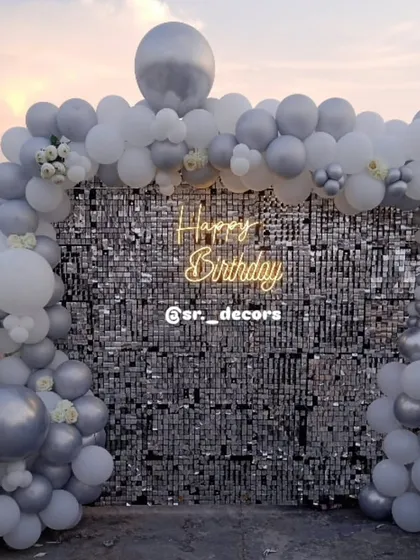 We take the party anywhere, even your rooftop. This setup features a brilliant silver shimmer wall framed by a white and silver balloon arch, creating a chic and elegant photo corner for an outdoor celebration.