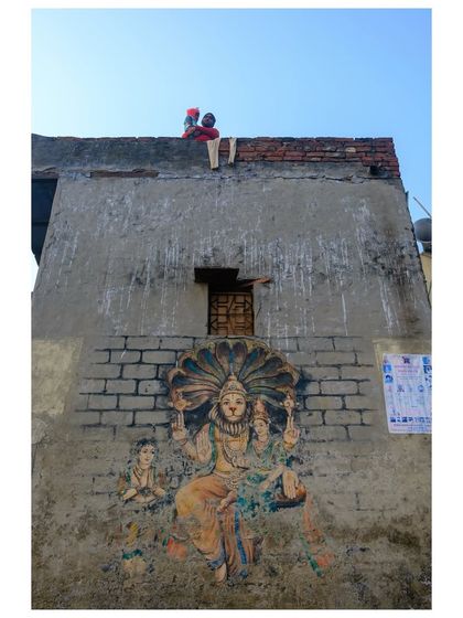 A view of a weathered building near the Yamuna, with a faded mural of a deity on its wall. On the roof above, two children are visible, adding a touch of life to the scene of urban decay.