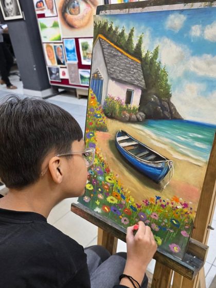 Another angle of the young artist at his easel. He is using a reference photo to guide his drawing of the boat and the shoreline.