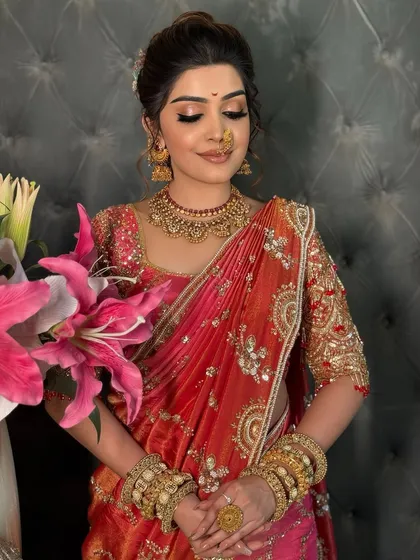 A bride in a pink and orange saree, holding flowers and looking serene.