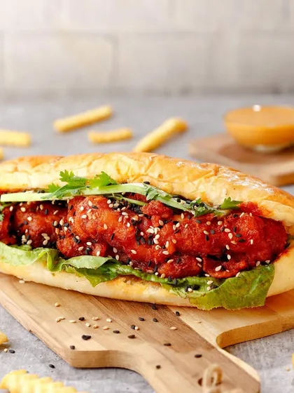 A loaded Korean-style chicken sandwich on a wooden board, surrounded by scattered fries.