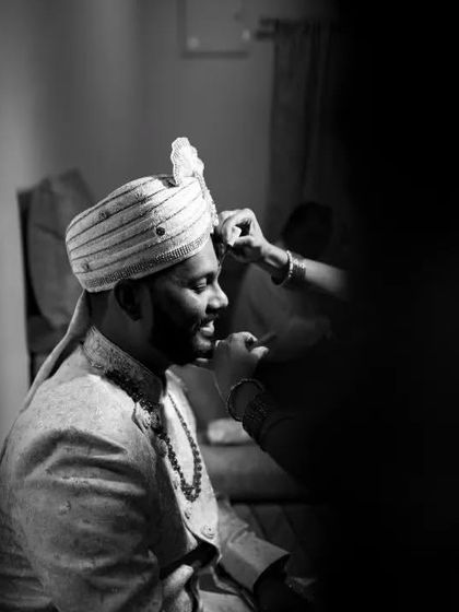 A candid black and white shot of the groom getting his turban adjusted, a quiet moment of preparation before the main ceremony.