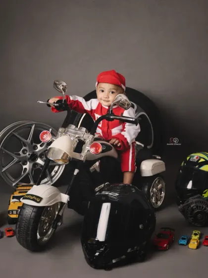 A toddler enjoying his biker-themed photoshoot, sitting on a miniature motorcycle. The set is complete with helmets, wheels, and toy cars for an authentic motor-themed portrait.