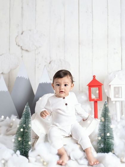 A cozy 'Winter Wonderland' themed session. This little one is dressed in a simple white outfit, sitting amongst fake snow, miniature pine trees, and mountain cutouts.