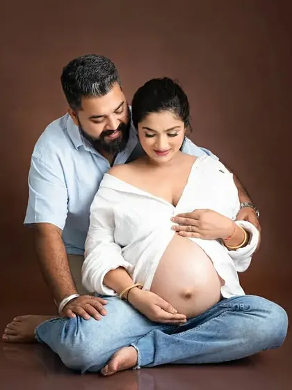 A relaxed, seated portrait of an expecting couple during a studio maternity session. The brown backdrop provides a warm and intimate feel for casual poses.