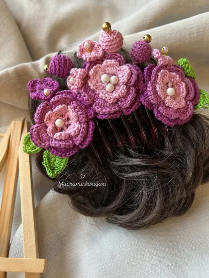 A close-up of the purple floral crochet bun comb, showing the intricate details of the petals and the pearl accents. It's designed to sit beautifully on a hair bun.