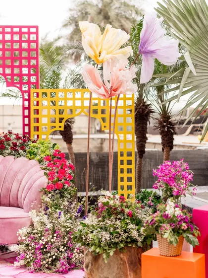 A Haldi setup with a vibrant, eclectic feel. The decor includes a pink scalloped sofa, colorful lattice backdrops, and oversized paper flowers, all surrounded by lush greenery.