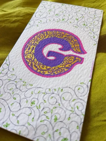 An illuminated letter 'G' decorated with intricate gold details and delicate vine-like flourishes. This style of work is perfect for creating custom monograms for wedding invitations or personalized stationery.