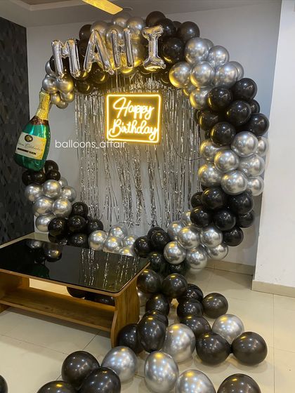 A chic birthday party with a black and silver theme. The circular balloon arch is decorated with a champagne bottle foil balloon and a 'Happy Birthday' neon sign.