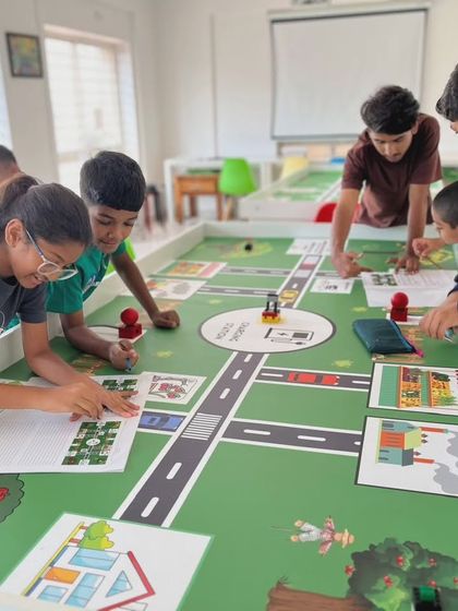 A group of students works together on the IRO 2024 challenge mat, discussing strategy and planning their robot's next move to efficiently manage the farm-themed tasks.