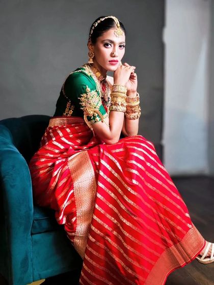 A South Indian inspired bridal look for an editorial feature. The hair is styled in a classic bun adorned with a simple gold hair accessory, perfectly complementing the vibrant red and gold silk saree.