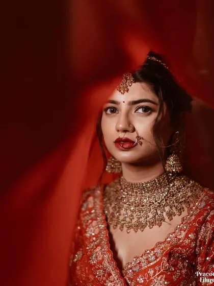 A striking bridal portrait with a bold red background. The bride's direct gaze and stunning gold jewelry create a powerful and captivating image.