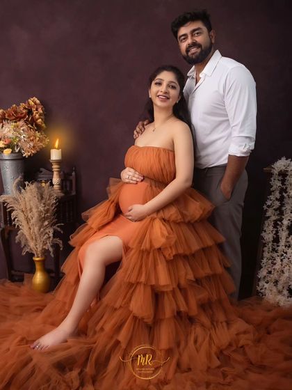 A happy and relaxed couple's portrait. The mother-to-be is seated in a vibrant orange gown, with her partner standing beside her in a rustic, candlelit studio setting.