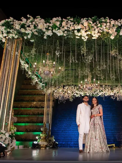 The couple posing for a photo under their magnificent wedding stage, adorned with a canopy of white flowers and hanging chandeliers.