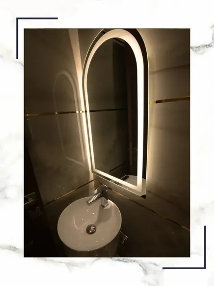 An arched, backlit mirror serves as the stunning centerpiece in this bathroom, complemented by a minimalist pedestal sink and sleek fixtures.