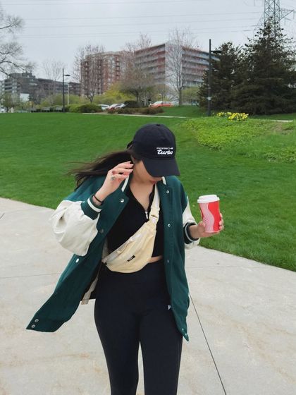 This candid, on-the-move shot shows a casual, sporty outfit in a park setting. It features a varsity jacket, leggings, and a coffee cup, creating a relatable "day in the life" moment.