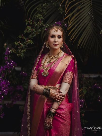A full-length portrait of a bride in a gorgeous pink saree, her arms adorned with dark, stained henna.