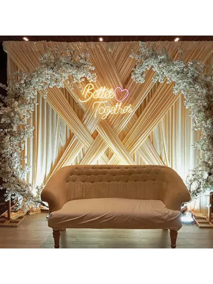 An engagement party stage featuring crisscross draping in a soft champagne color. The look is completed with white cherry blossom floral arches and a 'Better Together' neon sign for a modern, romantic feel.