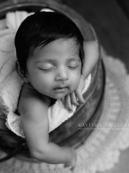 A two-month-old baby sleeps in a wooden bucket, captured in timeless black and white. The angle and the baby's slight smirk create a beautiful and artistic portrait.