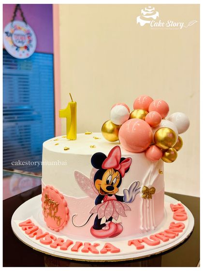 A sweet Minnie Mouse cake for a first birthday. This design features a fairy Minnie illustration, a plaque with the baby's name, and a cascade of pink and gold balloons.