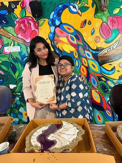 A participant and instructor pose with a certificate in front of a unique purple and white geode resin clock, celebrating a successful workshop.