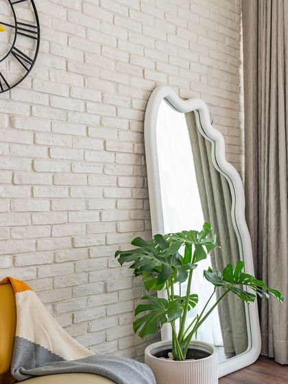 A cozy reading nook in the 'Urban Oasis' bedroom. The wavy floor mirror, monstera plant, and comfortable armchair create a perfect spot to relax next to the textured brick wall.