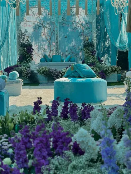 A view of the 'Firozi Ishq' setup through a field of purple and white larkspur. The color contrast between the flowers and the turquoise decor creates a visually stunning and serene atmosphere.