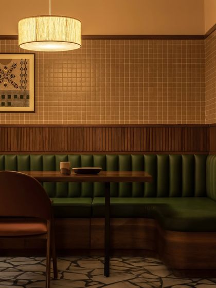 This cozy booth at the Grid Pavilion restaurant offers an intimate dining setting. The space is defined by rich textures, from the vertically stitched green upholstery to the ribbed wooden wall paneling and the warm glow of a custom fabric-wrapped pendant light.