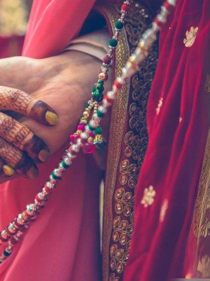 A close-up detail shot of hands during a wedding ritual. The colorful beads and intricate henna patterns highlight the rich textures and traditions of an Indian wedding.