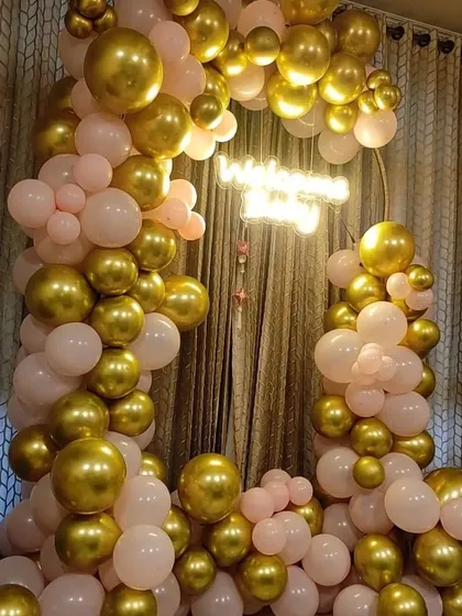 A soft and elegant balloon arch in pink and gold. This setup is perfect for birthdays or anniversaries, featuring a 'Happy Anniversary' neon sign nestled within the balloons.