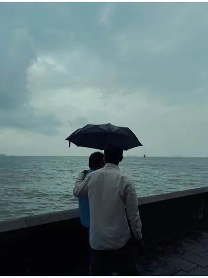 A couple stands close under an umbrella, looking out at the sea from the Marine Drive promenade. The composition is simple and intimate, focusing on their connection against the vast, grey water.