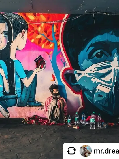 A poignant image of a homeless man sitting in front of a mural that depicts themes of hope, nature, and the pandemic. The real-life scene adds a powerful layer of meaning to the artwork behind it.