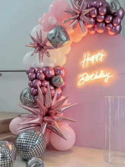 A trendy disco-themed birthday decoration. This setup features a pink arched backdrop, a neon sign, and a garland of pink and chrome balloons mixed with starburst and disco ball foils.