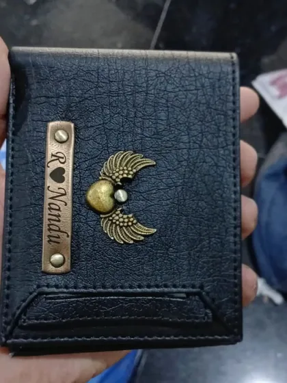 A personalized black wallet with a heart and wings charm, held to show the texture and craftsmanship.